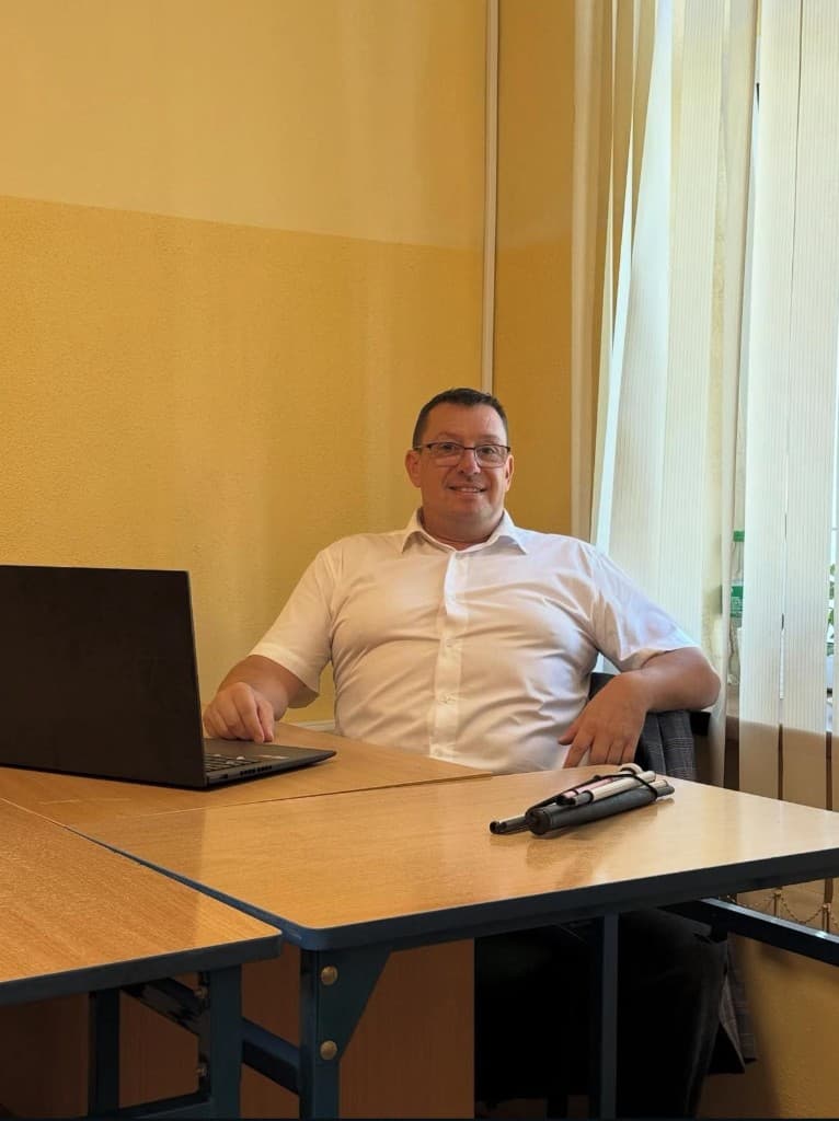 Sebastian, katecheta — przy biurku w szkole; na biurku między innymi laptop i biała laska